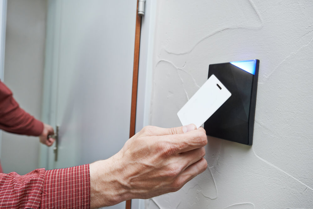 Mão aproximando cartão de RFID branco de leitora preta com indicador luminoso azul instalada ao lado de porta