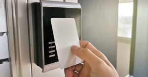 Mão aproximando cartão branco de proximidade de leitora preta com botões numéricos instalada em porta
