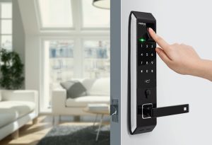 Fechadura biométrica Intelbras preta instalada em porta, com teclado numérico, leitor de digital e maçaneta, em sala com sofás claros ao fundo