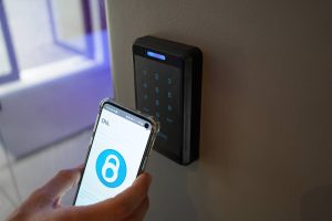 Mão segurando smartphone com aplicativo de desbloqueio próximo a leitor de controle de acesso com teclado numérico iluminado em azul