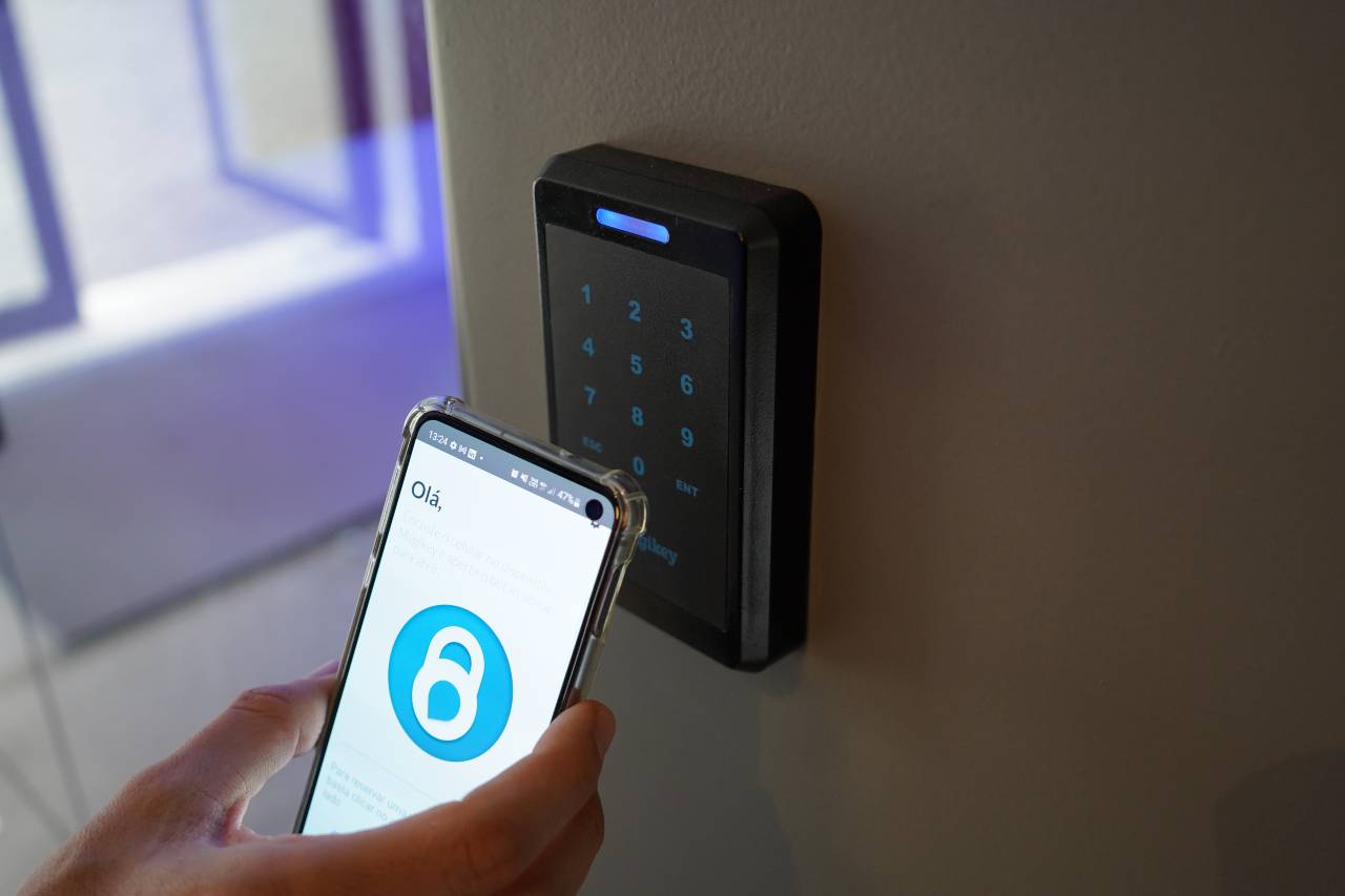 Mão segurando smartphone com aplicativo de desbloqueio próximo a leitor de controle de acesso com teclado numérico iluminado em azul