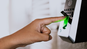 Close-up de dedo posicionado sobre leitor biométrico com luz verde de leitura válida, instalado próximo a uma porta