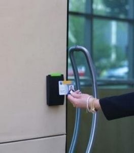Mão aproximando cartão de proximidade de leitor instalado ao lado de porta de vidro com maçaneta metálica