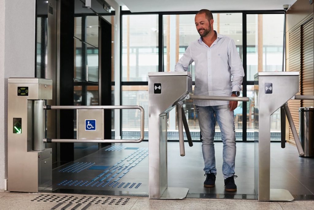 Homem passando por catraca tripé em aço inox com leitor de proximidade em recepção corporativa moderna, ao lado de portão acessível