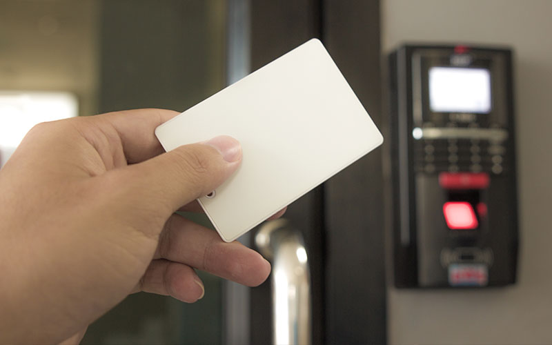 Mão segurando cartão branco próximo a terminal de controle de acesso com tela, teclado e leitor biométrico com luz vermelha