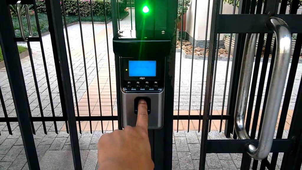 Dedo pressionando leitor biométrico com tela azul e indicador luminoso verde, instalado em portão de condomínio