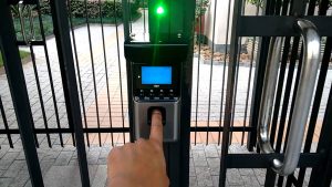 Dedo pressionando leitor biométrico com tela azul e indicador luminoso verde, instalado em portão de condomínio